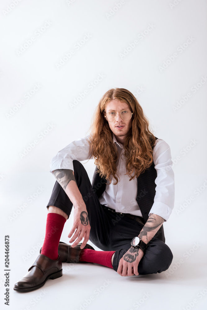 Fototapeta premium stylish tattooed businessman with curly hair sitting on floor and looking at camera on white
