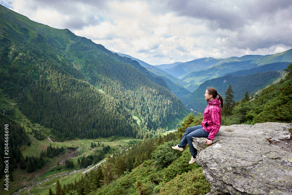 Smiling beautiful female tourist sitting on rock edge admiring breathtaking view of green grassy slopes and mountains with trees, fir trees and pines in Romania. Woman climber happy amazing nature