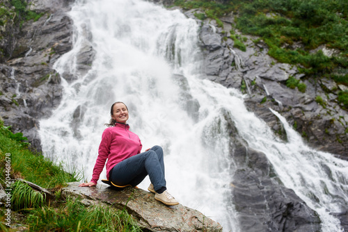 Happy woman smiling to the camera sitting in front of a beautiful Balea waterfall in Fagarash mountains copyspace power nature health happiness lifestyle activity camping hiking sports.