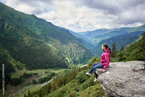 Smiling beautiful female tourist sitting on rock edge admiring breathtaking view of green grassy slopes and mountains with trees, fir trees and pines in Romania. Woman climber happy amazing nature