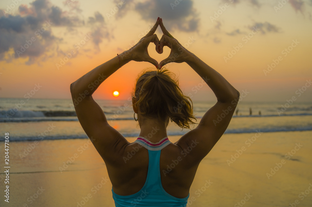 back portrait of young fit and healthy attractive woman doing love heart shape with hands and fingers against amazing beautiful sunset beach in yoga