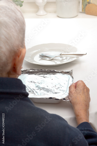 Old woman sitting alone at a table with plate, cutlery and a precooked meal from a food delivery service.