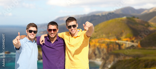 travel, tourism and male friendship concept - group of friends hugging over bixby creek bridge on big sur coast of california background