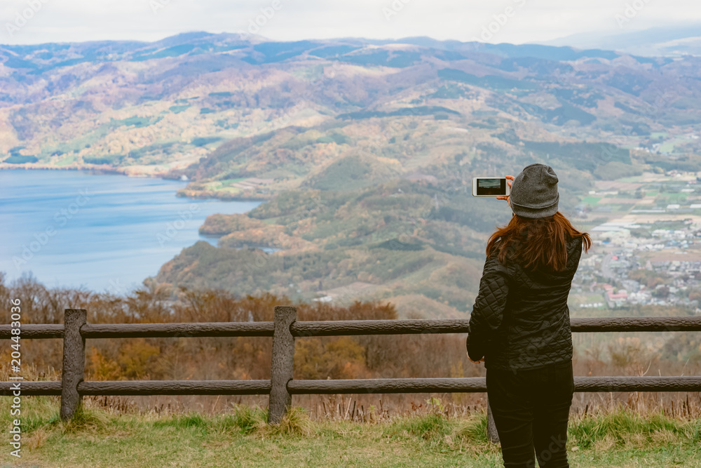 A female tourist using her smartphone to capture the beautiful landscape view of Lake Toya, Hokkaido, Japan.