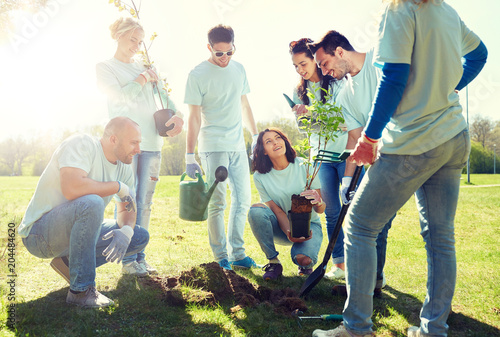 volunteering, charity, people and ecology concept - group of happy volunteers planting tree and digging hole with shovel in park