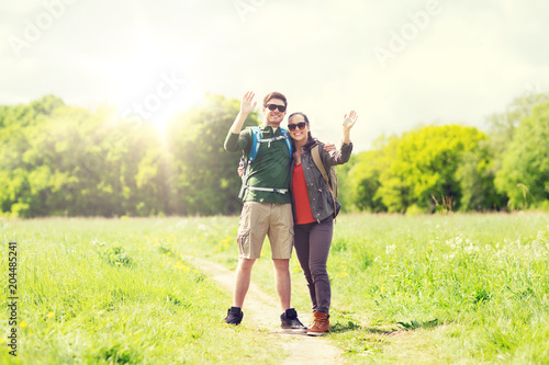 travel, hiking, backpacking, tourism and people concept - happy couple with backpacks waving hands and walking along country road outdoors