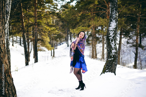 stylish brunette woman with dark hair in a blue dress posing in the open frosty air in a forest among the trees in a traditional Russian wool shawl