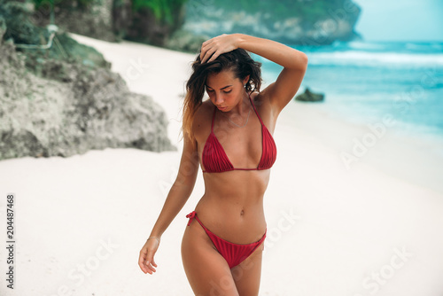 Young girl with a gorgeous body is resting on the beach with white sand near the ocean. Beautiful sexy model in a red bathing suit sunbathing. Brunette with curly hair in bikini.