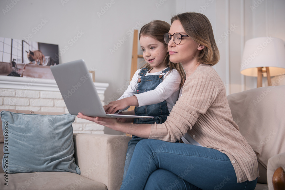 Freelancing. Beautiful concentrated mother sitting with her daughter and working on the laptop Freelancing. Beautiful concentrated mother sitting with her daughter and working on the laptop