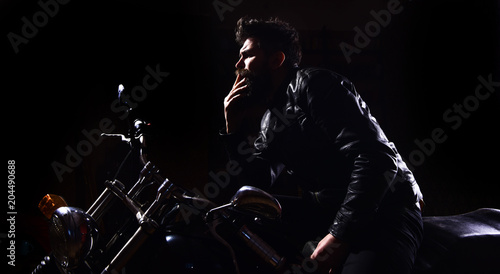 Night racer concept. Man with beard, biker in leather jacket sitting on motor bike in darkness, black background. Macho, brutal biker in leather jacket riding motorcycle at night time, copy space.