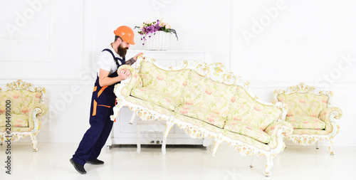 Loader moves sofa  couch. Courier delivers furniture in case of move out  relocation. Delivery service concept. Man with beard  worker in overalls and helmet lifts up sofa  white background.