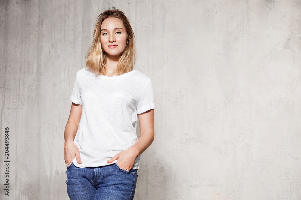 Portrait of cheerful girl posing at studio. Lovely woman standing with hands in pockets wearing in white jersey and jeans. She is looking at camera with happiness. Copy space in right side Portrait of cheerful girl posing at studio. Lovely woman standing with hands in pockets wearing in white jersey and jeans. She is looking at camera with happiness. Copy space in right side