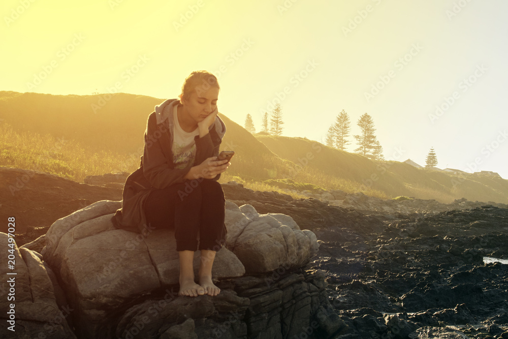 young woman sitting on some rocks at the beach reading her cellphone