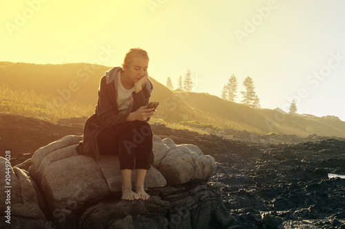 young woman sitting on some rocks at the beach reading her cellphone