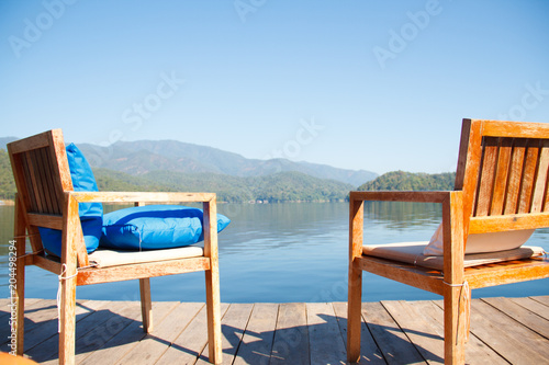 Two wooden chairs on terrace near the lake with blurred mountain and blue sky natural background. From behind.