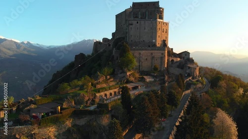 Aerial: flying around old medieval abbey perched on mountain top, background snowy Alps at sunrise. Sacra di San Michele (italian) - Saint Michel Abbey (english traslation) - Turin, Italy  photo