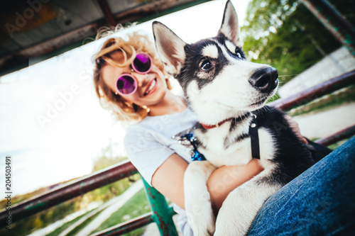 blonde with curly hair in pink glasses sits on the bench with husky on her hands in the spring in the park