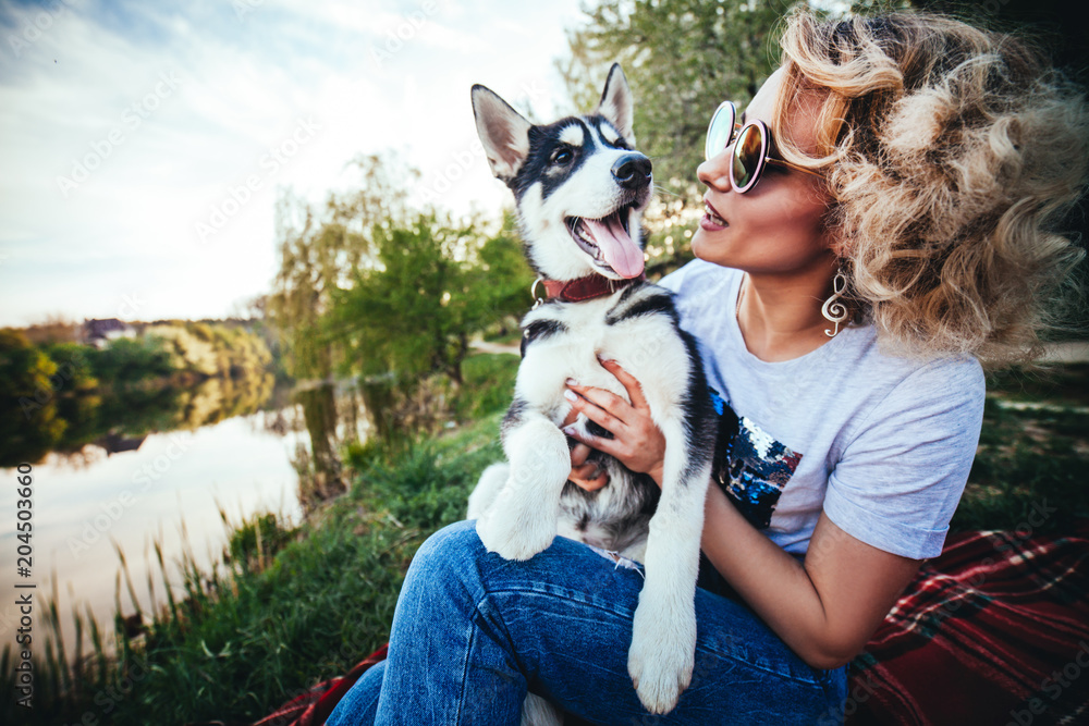 blonde in glasses and with earrings in the form of a treble clef smiles and looks at a small Husky with an open mouth near the river in the summer blonde in glasses and with earrings in the form of a treble clef smiles and looks at a small Husky with an open mouth near the river in the summer