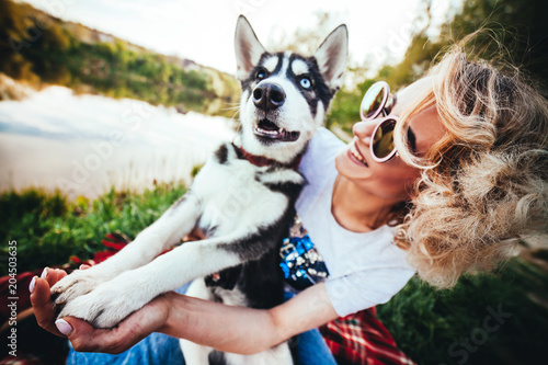 joyful girl in pink glasses is holding in her hands and looking on small husky with different eyes color near the river in the summer