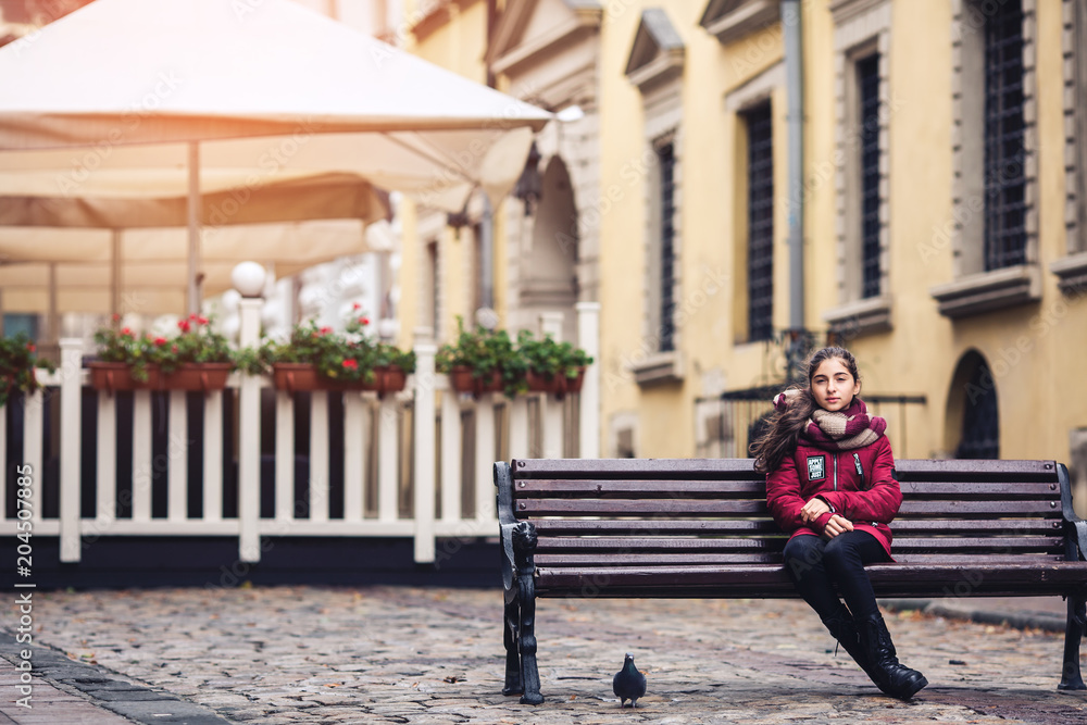 Beautiful autumn girl sitting on bench in middle of street on cafe background
