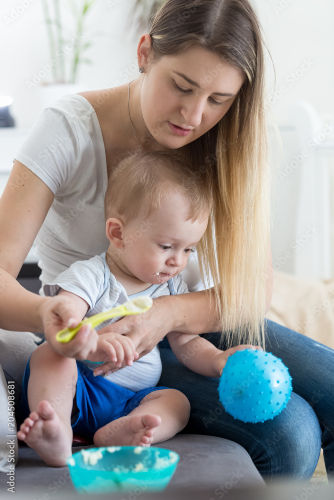 Young mother sitting on sofa with her baby son and feeding him from spoon Young mother sitting on sofa with her baby son and feeding him from spoon