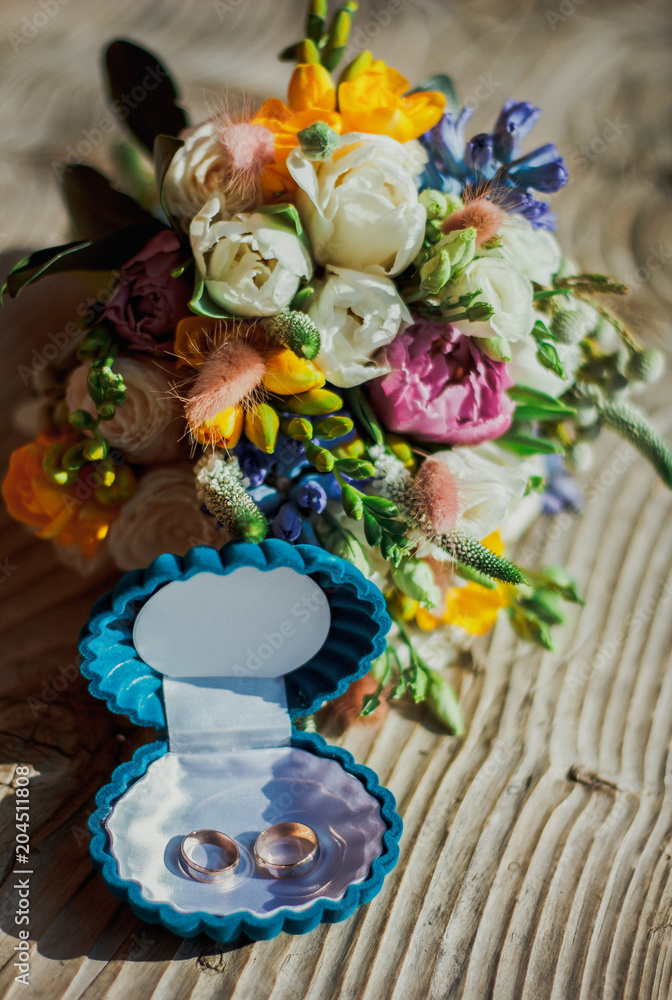 Bright summer wedding bouquet is lying on the wooden bench. Sun lights on wedding decorative details.Wedding rings in blue box are nearby.