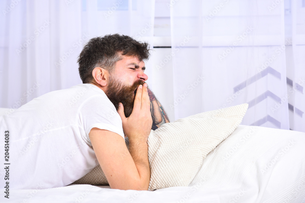 Man in shirt yawning while laying on bed, white wall and curtain on background. Guy on sleepy tired face yawning. Macho with beard and mustache yawning, relaxing, having nap, rest. Sleepyhead concept.