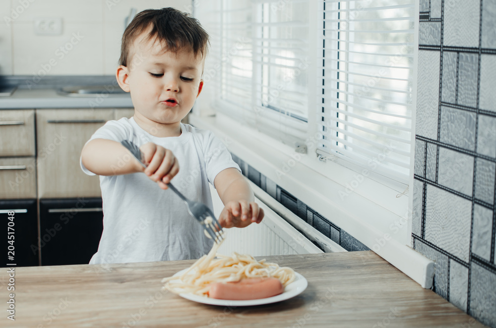 baby eating pasta with a fork and sausages in a white t-shirt in the kitchen