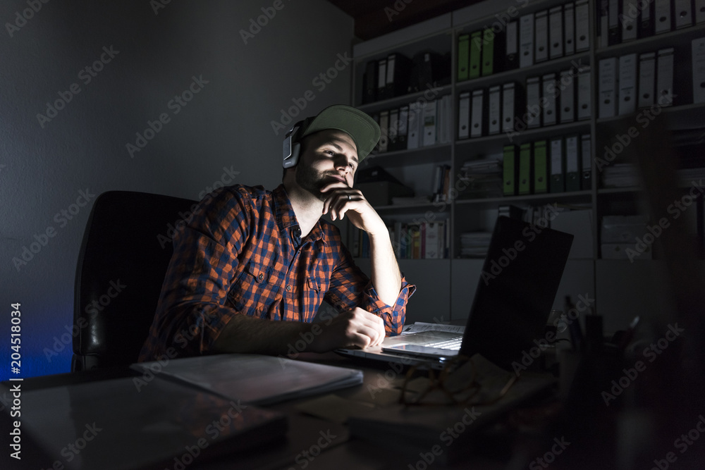 Portrait of freelancer sitting at desk at night using laptop and headphones Portrait of freelancer sitting at desk at night using laptop and headphones