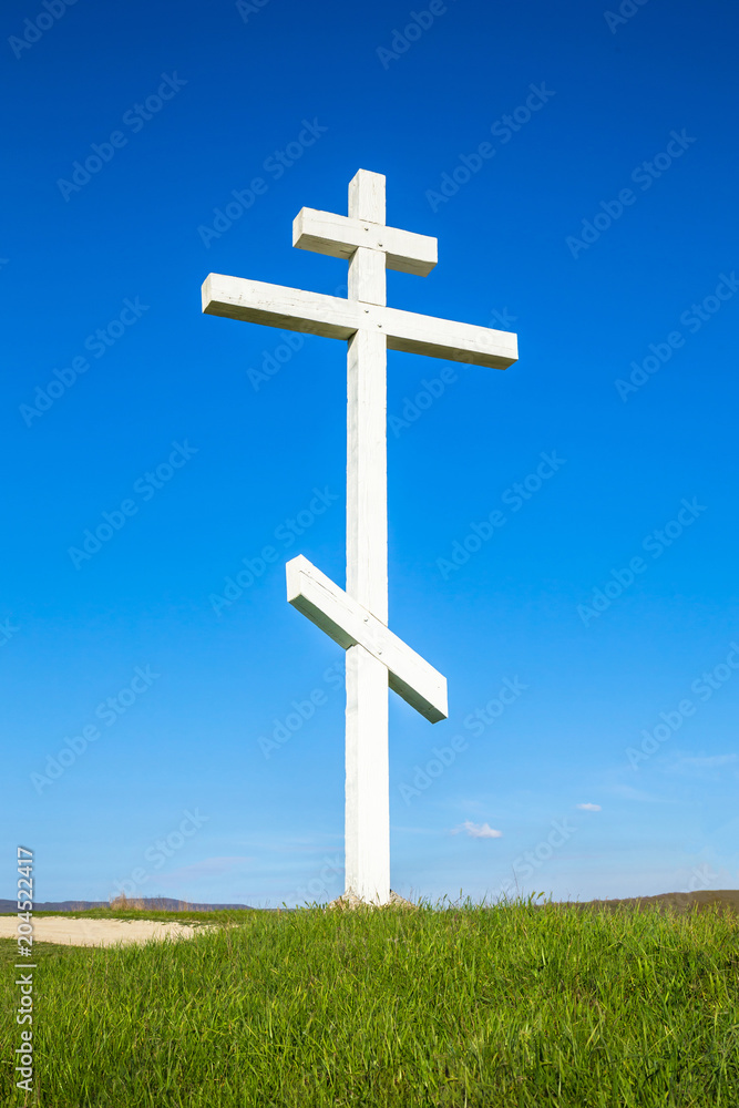 A wooden cross on top of a hill. Orthodox white cross glows on the top of the hill on the background of blue sky and of green grass