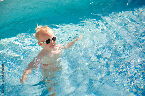 Cute blonde little baby with sunglasses splashing happily in the pool with clear blue water in the summer. Top view. Copy space.