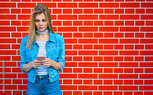 Young pretty woman type message - Female jeans fashion portrait - Teenager student holding mobile phone for sms next to brick wall background