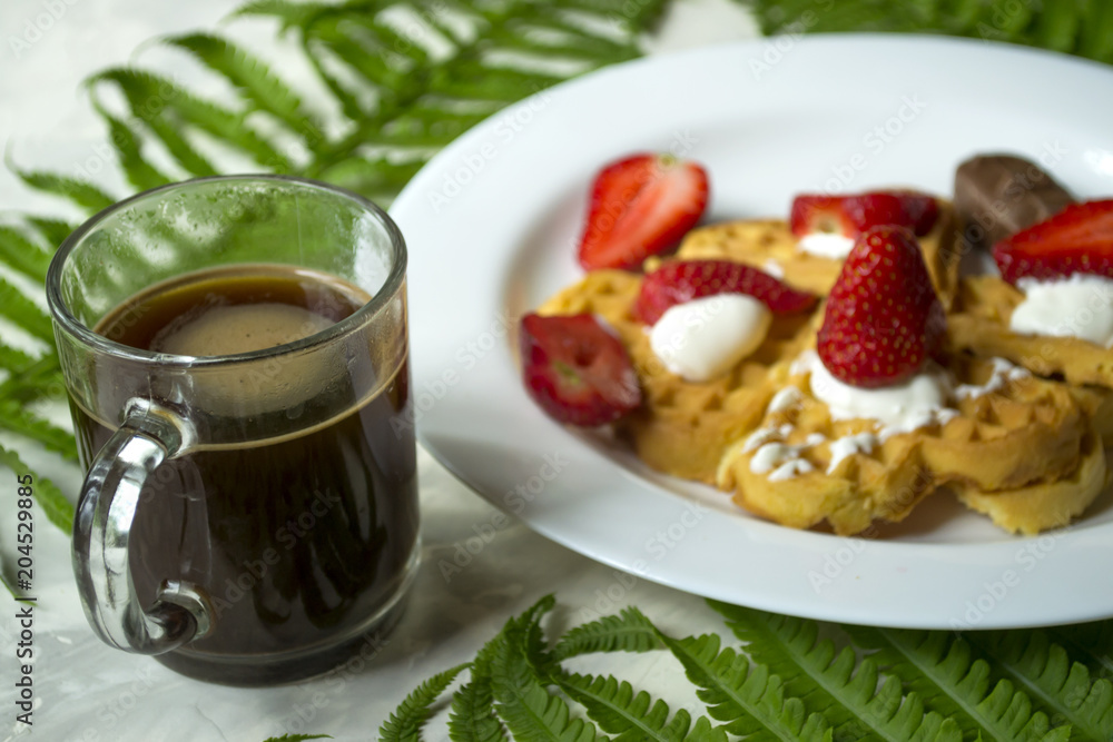 Waffles with strawberry, a cup of coffee and green leaves of fern on a table. Beautiful and tasty breakfast. Waffles with strawberry, a cup of coffee and green leaves of fern on a table. Beautiful and tasty breakfast.