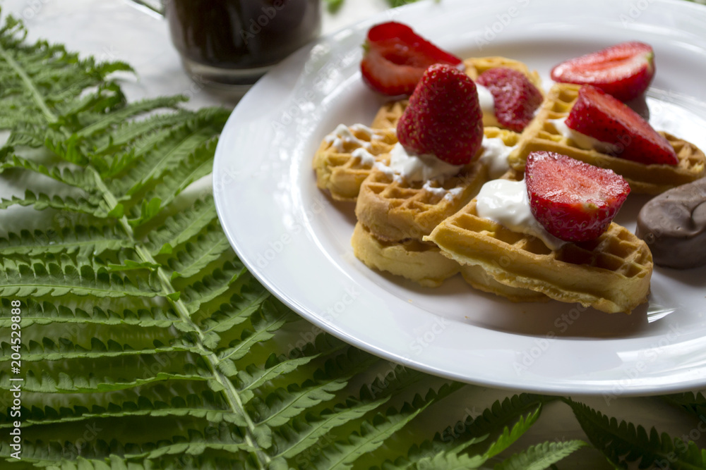 Waffles with strawberry on a plate, green leaves of fern on a table. Close up. Beautiful and tasty breakfast.