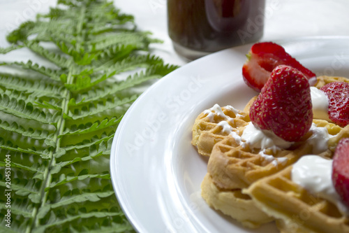 Waffles with strawberry on a plate, green leaves of fern on a table. Close up. Beautiful and tasty breakfast.