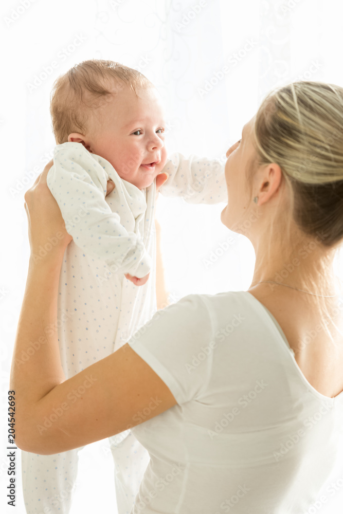 Happy young woman holding her smiling 3months old baby on stretched hands at window Happy young woman holding her smiling 3months old baby on stretched hands at window