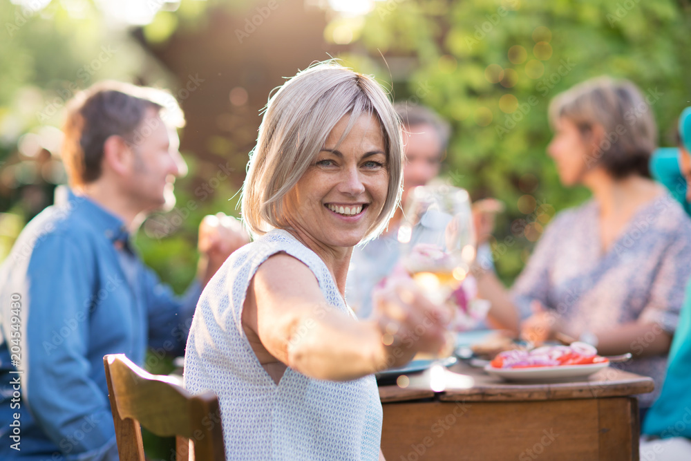 In summer. a group of friends in their forties gathered around a table in the garden to share a meal. They toast with their glasses of wine to the camera.