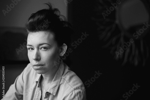 Closeup black and white portrait of beautiful sexy edgy young Caucasian woman. European female with dark hair sitting on sofa couch indoors. Androgen boyish stylized shoot. Film noise grain imitation