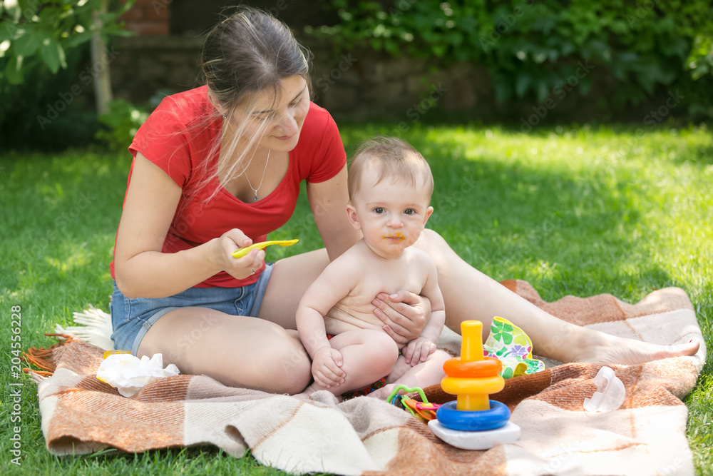 Young mother having family picnic with her baby boy and feeding him with porridge Young mother having family picnic with her baby boy and feeding him with porridge