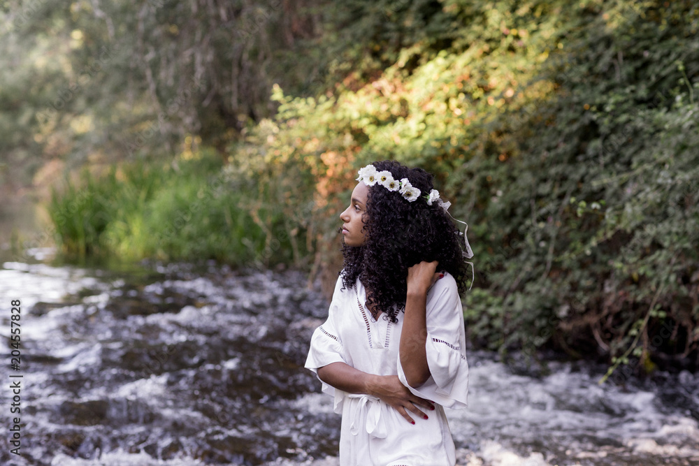 portrait outdoors of a beautiful young afro american woman at sunset. Green background. Lifestyle at nature. Wearing white casual summer dress. She is standing in a river. portrait outdoors of a beautiful young afro american woman at sunset. Green background. Lifestyle at nature. Wearing white casual summer dress. She is standing in a river.