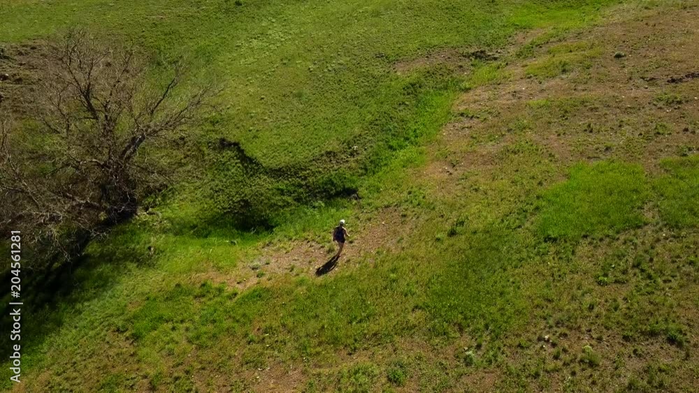 Aerial video of man walking in picturesque valley on green grass towards a river. Drone tracking shot of backpacker hiking in the mountains and hills. Hiker goes to the river. Aerial video of man walking in picturesque valley on green grass towards a river. Drone tracking shot of backpacker hiking in the mountains and hills. Hiker goes to the river.