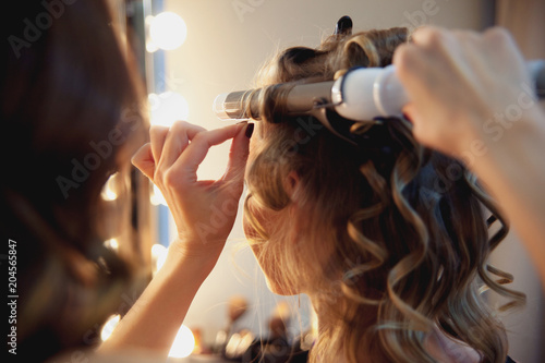 Hairstylist make beautiful curls with curler on light-brown with blonde client's hair. Creating a luxurious evening hairstyle with ringlets of medium length.