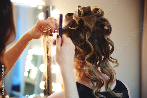 Hairstylist combs client's light-brown with blonde hair. Creating a luxurious evening hairstyle with ringlets of medium length.