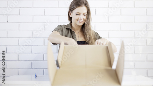 A smiling young woman opening cardboard box at home takes out a gift delivered gift by air mail