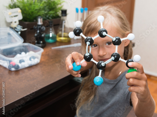 A little girl is at elementary school constructs a model of a molecule and conducts simple chemical experiments. A microscope, test tubes, a metal robot are on a table. STEM education. AI. 