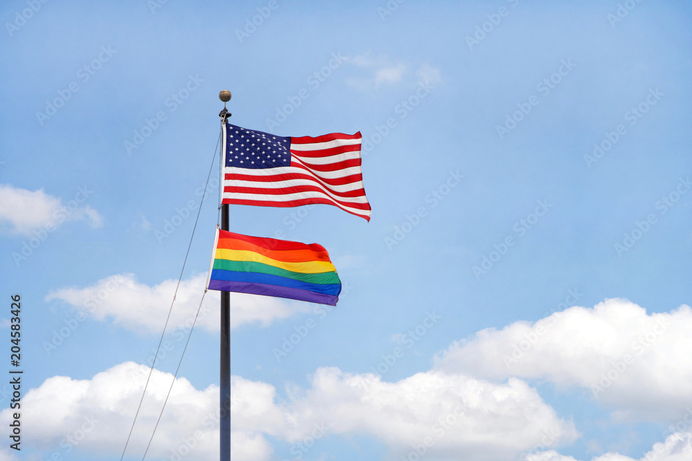 U.S. Flag flying in a blue cloudy sky with rainbow flag below blowing in the wind. Fluffy white clouds in a blue sky background.