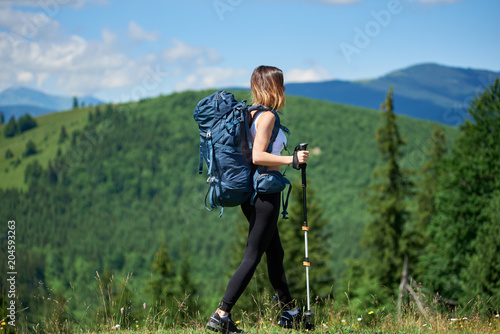Attractive sporty woman backpacker with blue backpack and trekking poles walking on the top of a hill, looking away, enjoying sunny day in the mountains. Concept of active lifestyle