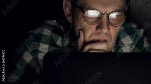 A man in glasses works late at night. He looks worried, he sees on the computer screen in front of him, a close-up
