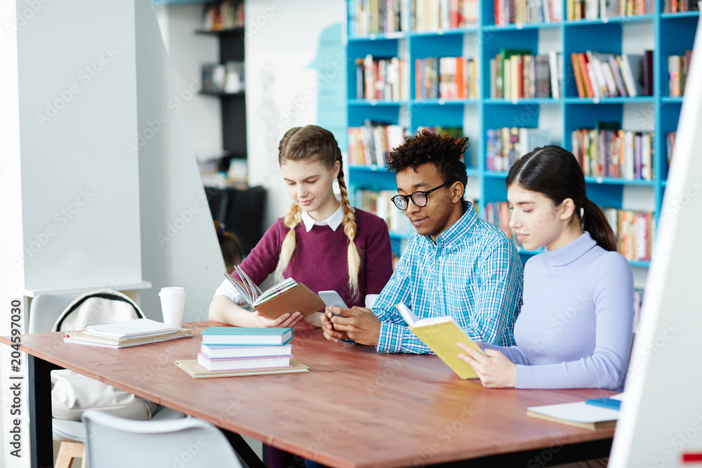 Two girls reading books in library while guy between them texting in smartphone after lessons Two girls reading books in library while guy between them texting in smartphone after lessons