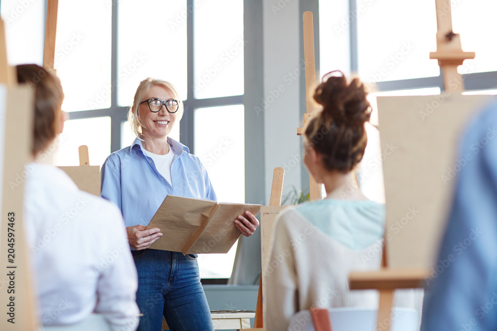 Successful teacher with open manual standing in front of her students and explaining them rules of professional painting Successful teacher with open manual standing in front of her students and explaining them rules of professional painting